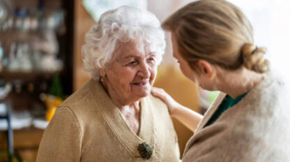 An elderly woman with white hair smiles at a person with blond hair, who gently touches her shoulder. This warm interaction reflects the compassionate environment often found in a mental health clinic.