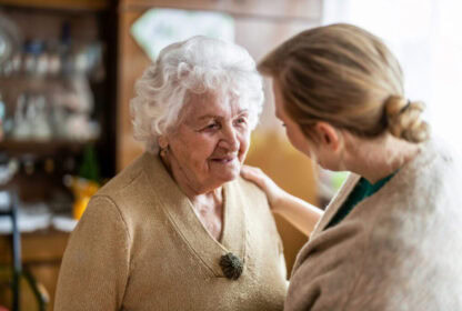 An elderly woman with white hair smiles at a person with blond hair, who gently touches her shoulder. This warm interaction reflects the compassionate environment often found in a mental health clinic.