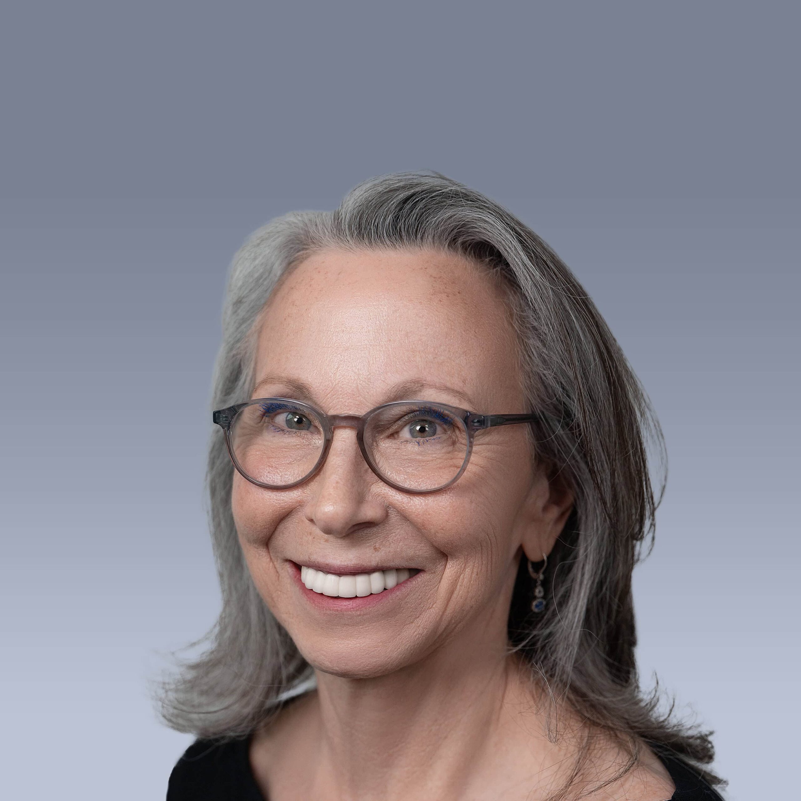 A smiling woman with gray hair and glasses against a gradient gray background. She is wearing a black top and earrings.