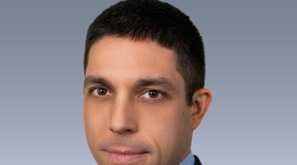 A headshot of a man with short dark hair and brown eyes, wearing a blue checkered shirt and a dark blazer, set against a plain grey background.