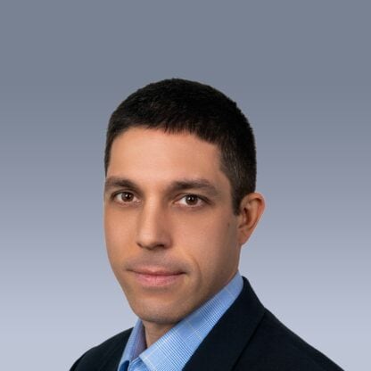 A headshot of a man with short dark hair and brown eyes, wearing a blue checkered shirt and a dark blazer, set against a plain grey background.