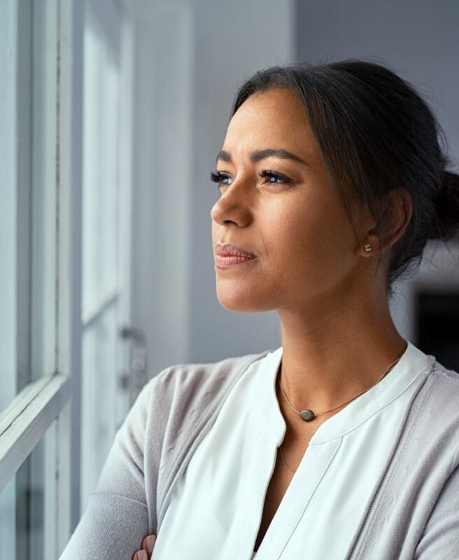 A woman with her hair in a bun stands indoors, looking thoughtfully out a window. She wears a light-colored cardigan and a white top, with a minimalist necklace. A wooden table is visible in the background.