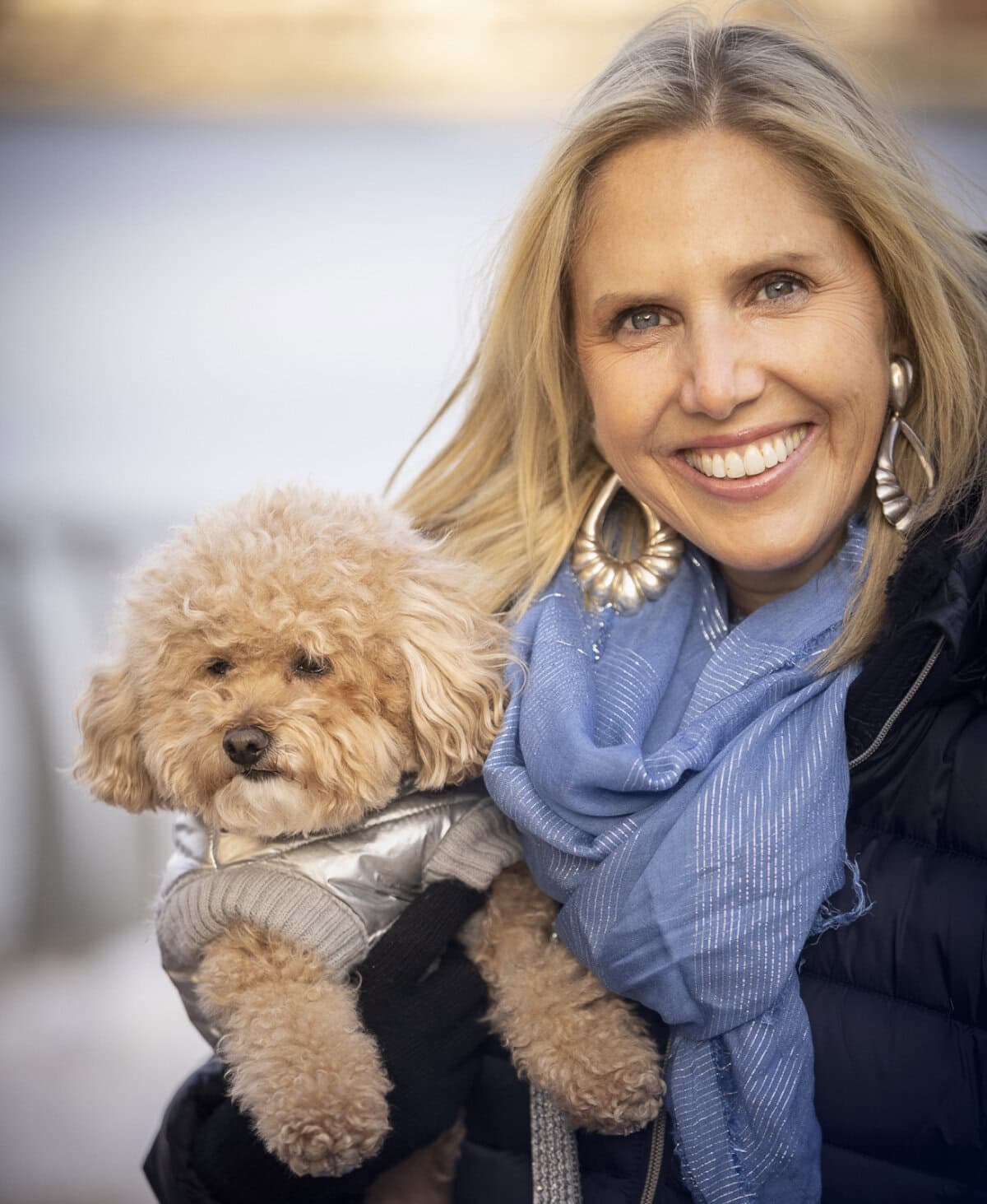 A woman smiles while holding a small, fluffy dog, offering solace akin to a gentle mental health treatment. She is wearing a blue scarf and earrings, and the dog has a gray sweater.
