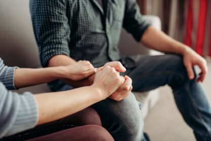 Two people sitting close, holding hands in a supportive manner, with one person wearing a checkered shirt and jeans, seated on a light-colored sofa.