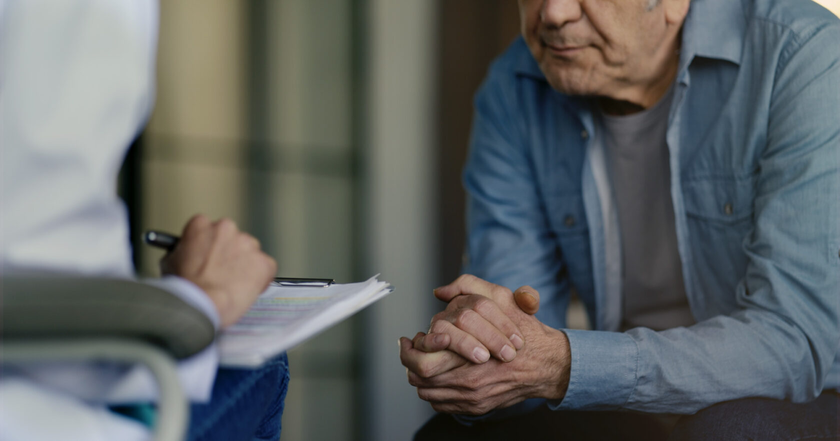 An older person seated, clasping their hands, conversing with a healthcare professional who is taking notes on a clipboard.