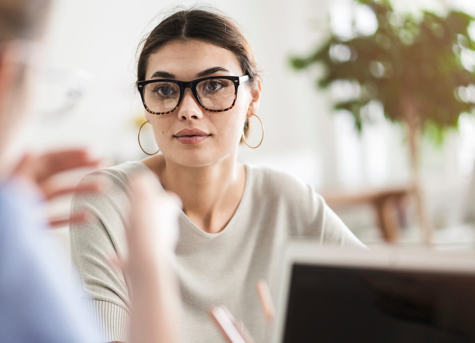 A person with glasses and hoop earrings sits attentively in a mental health clinic, looking towards another individual. They are indoors with a blurred background, creating an atmosphere focused on connection and care.