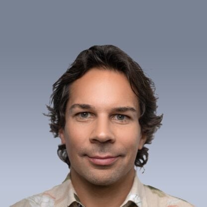 A person with shoulder-length dark hair and a slight smile looks directly at the camera against a gray background.