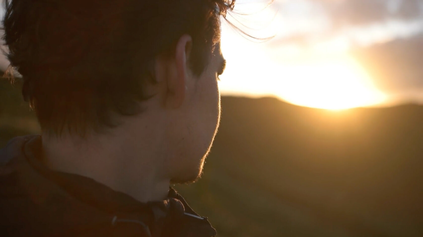 A person with short dark hair looks towards the setting sun over a mountainous landscape.