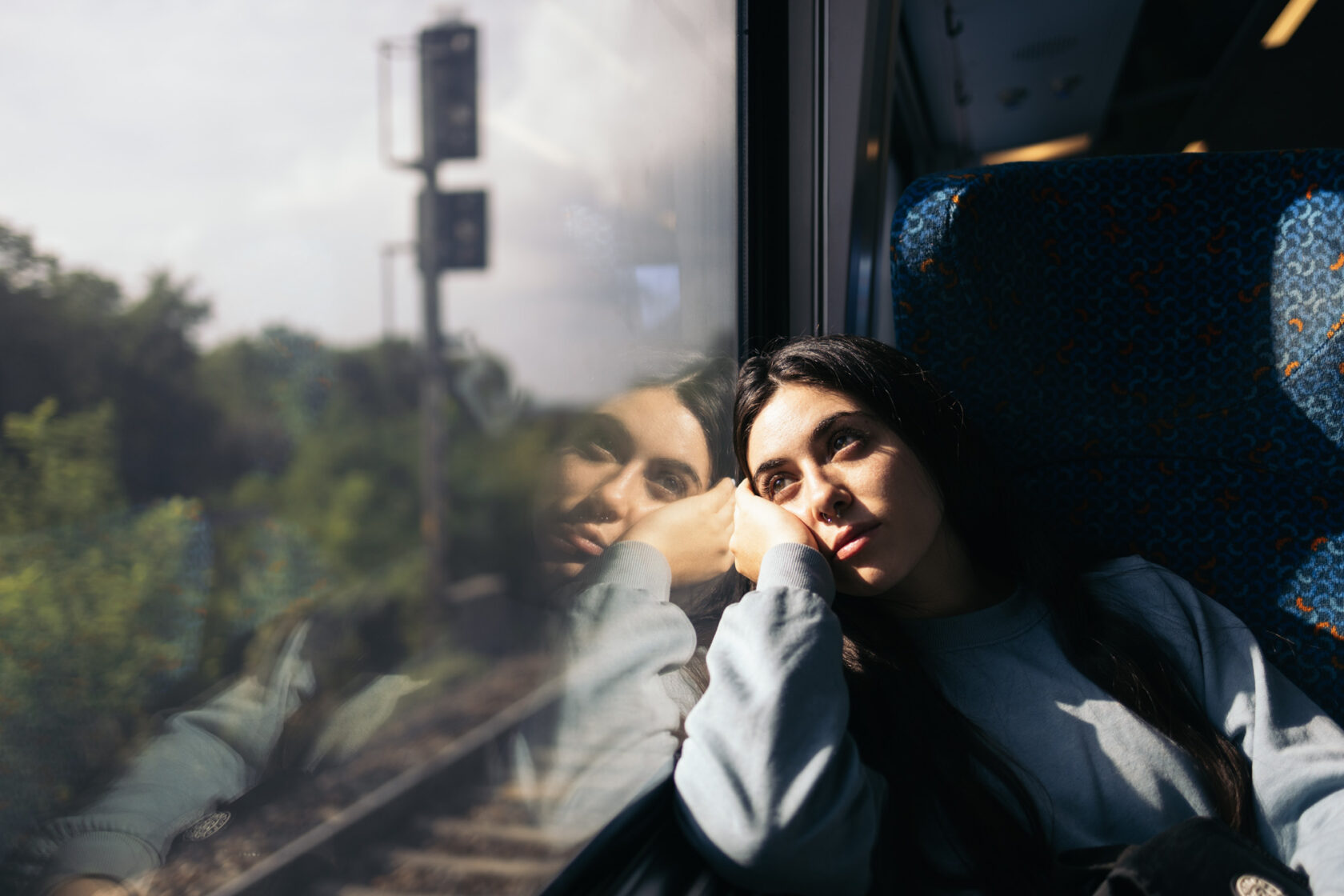 A person with long dark hair sits by a train window, resting their head on their hand, looking out at the scenery outside. Their reflection is visible in the window.