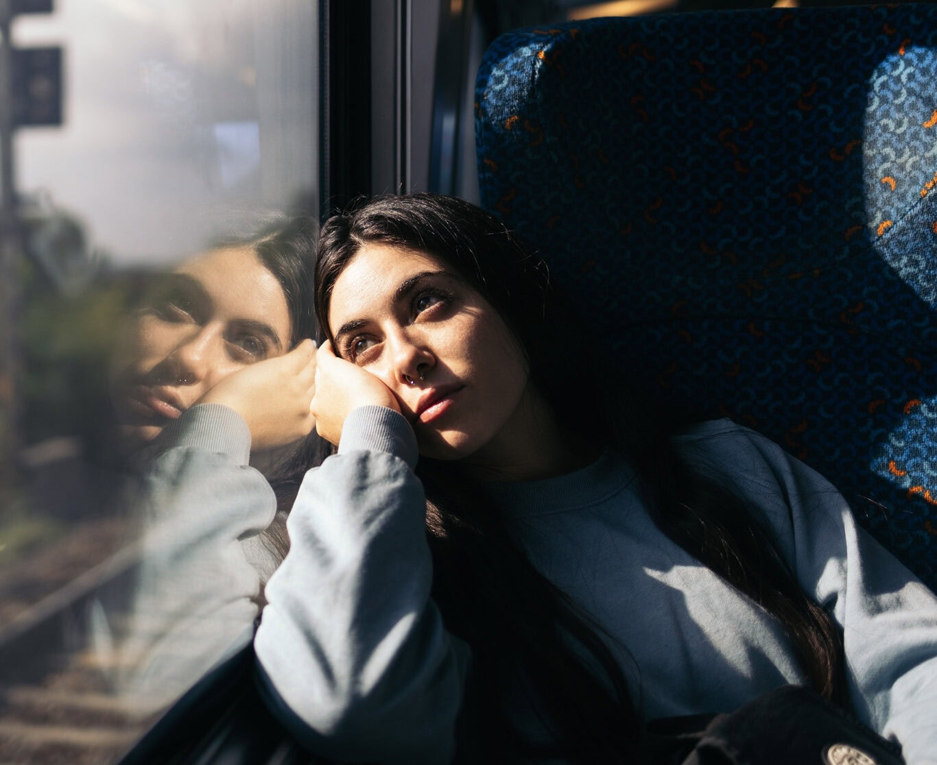 A person with long dark hair sits by a train window, resting their head on their hand, looking out at the scenery outside. Their reflection is visible in the window.