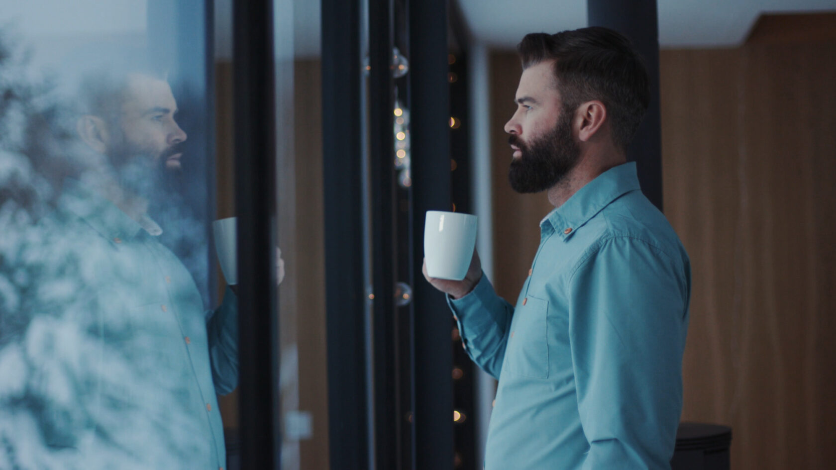 A man with a beard, wearing a teal shirt, holds a mug and looks out a window while his reflection appears on the glass.