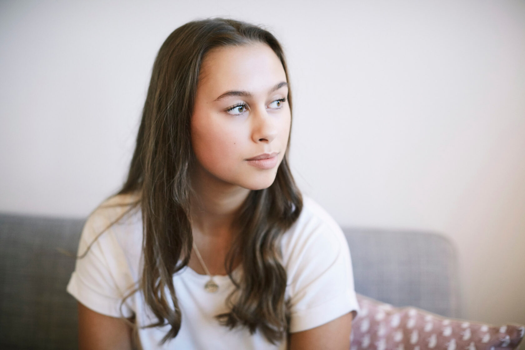 A young woman with long brown hair, wearing a white shirt, is sitting on a gray sofa. She is looking off to the side with a neutral expression.