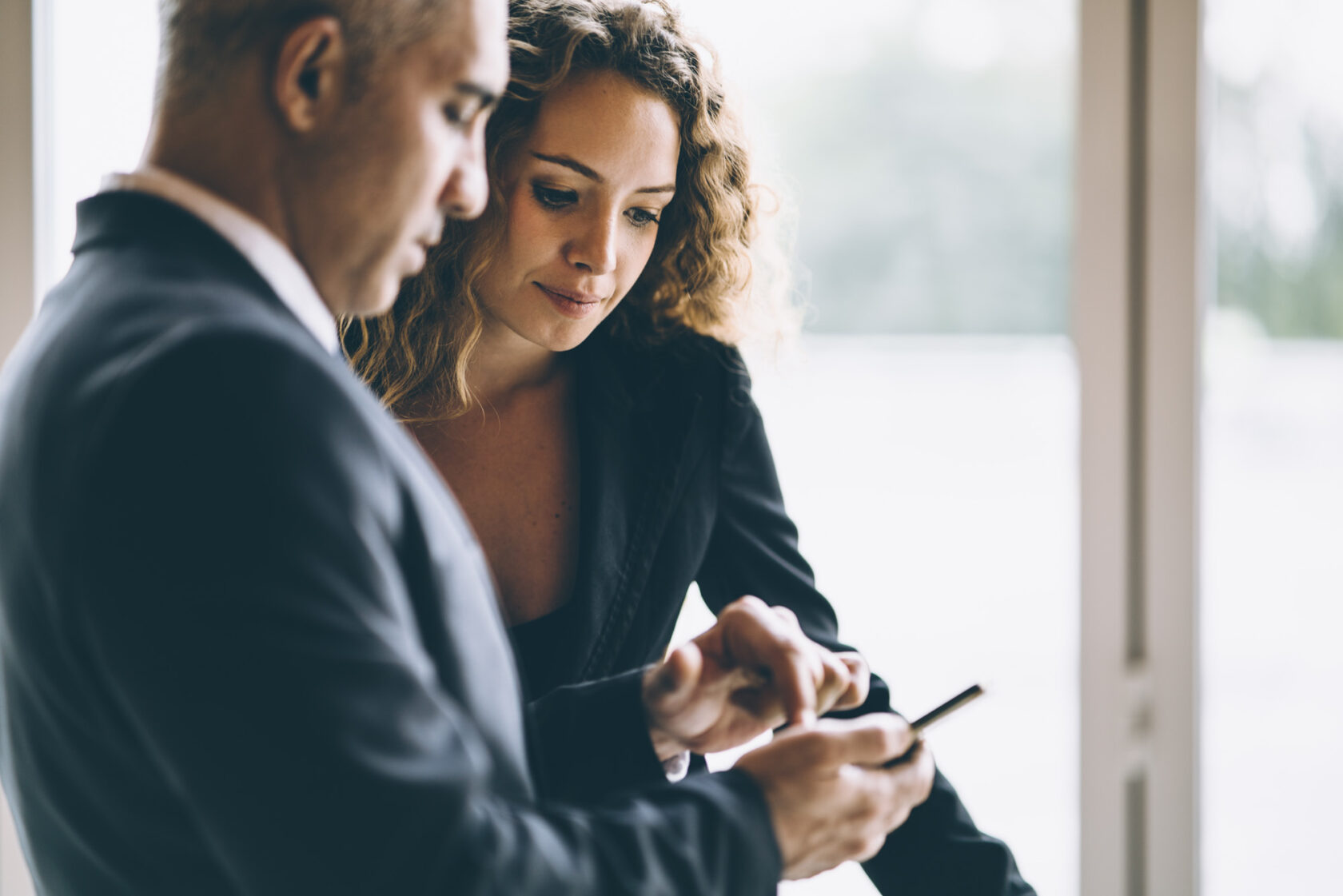 A man and a woman in business attire look at a smartphone together in an office setting.