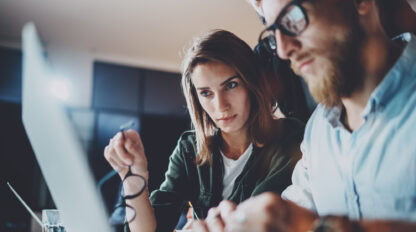 Two people are focused on a laptop screen. The woman on the left holds eyeglasses in her hand, while the man on the right, wearing glasses, types on the keyboard. A glass of water is in the background.