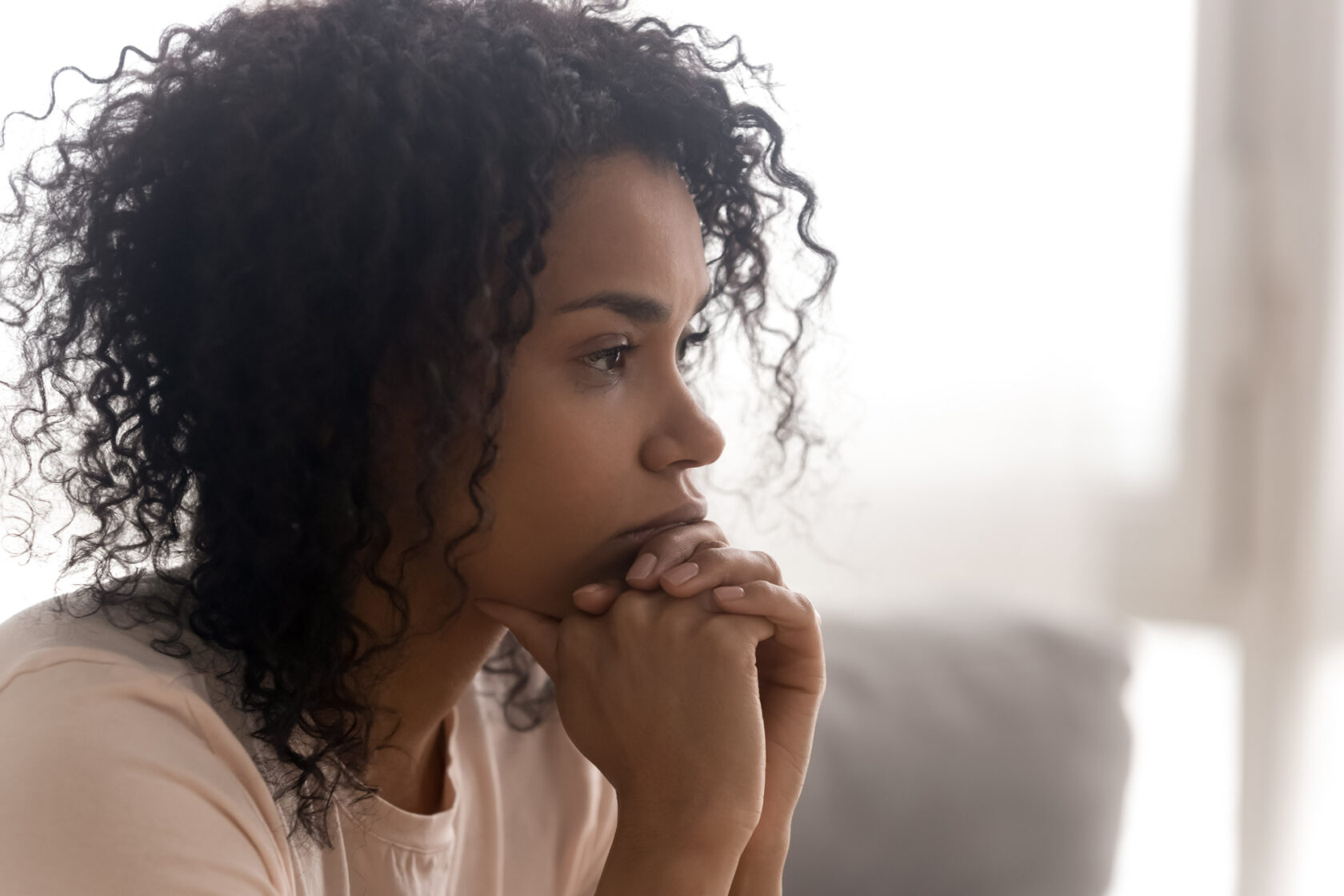 A woman with curly hair looks to the side, her face resting on her clasped hands, in a thoughtful expression.