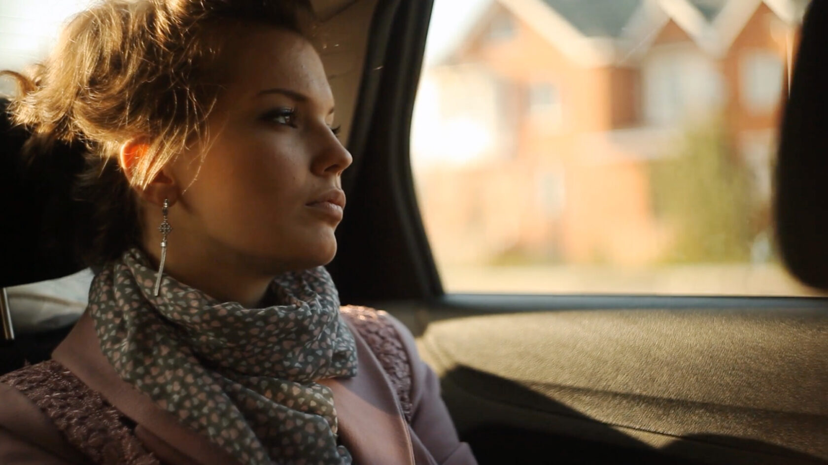 Woman with a polka dot scarf looks out the window of a car, with blurred houses visible in the background.