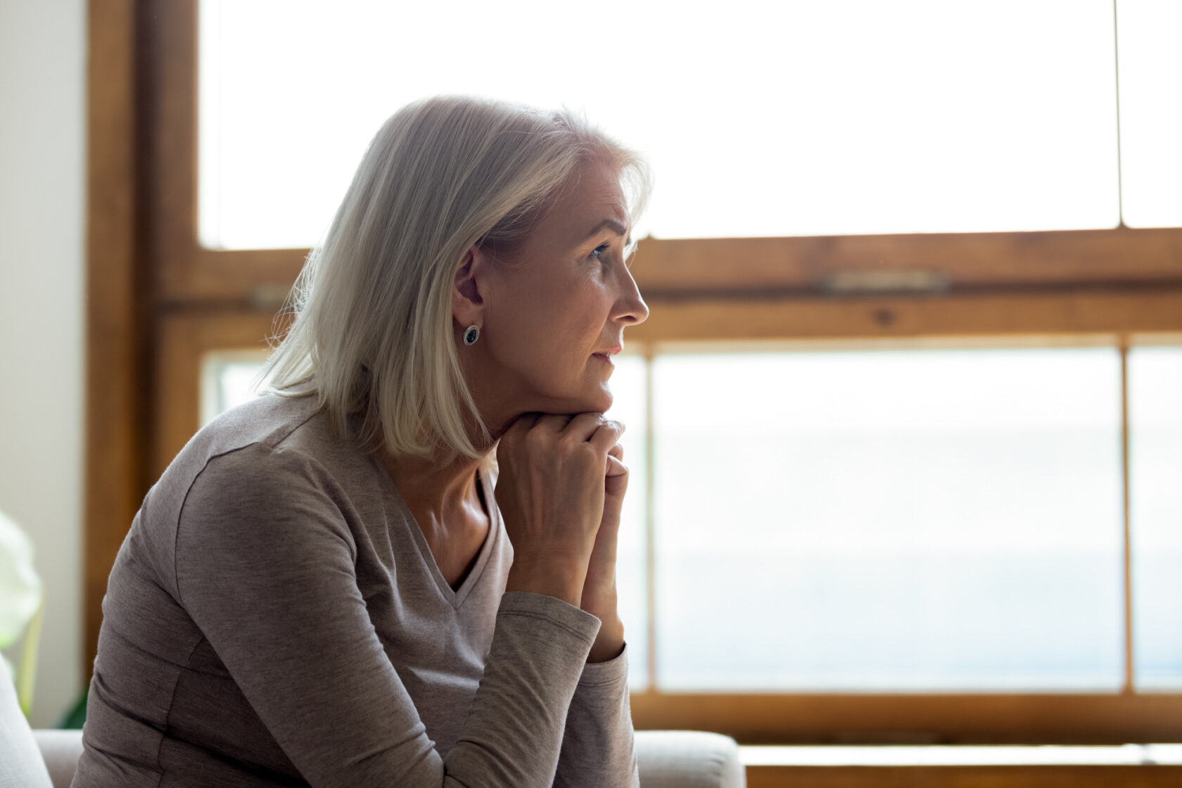 A woman with gray hair and a neutral expression, resting her chin on her hands, looks out of a window with wooden frames.