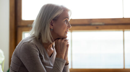 A woman with gray hair and a neutral expression, resting her chin on her hands, looks out of a window with wooden frames.