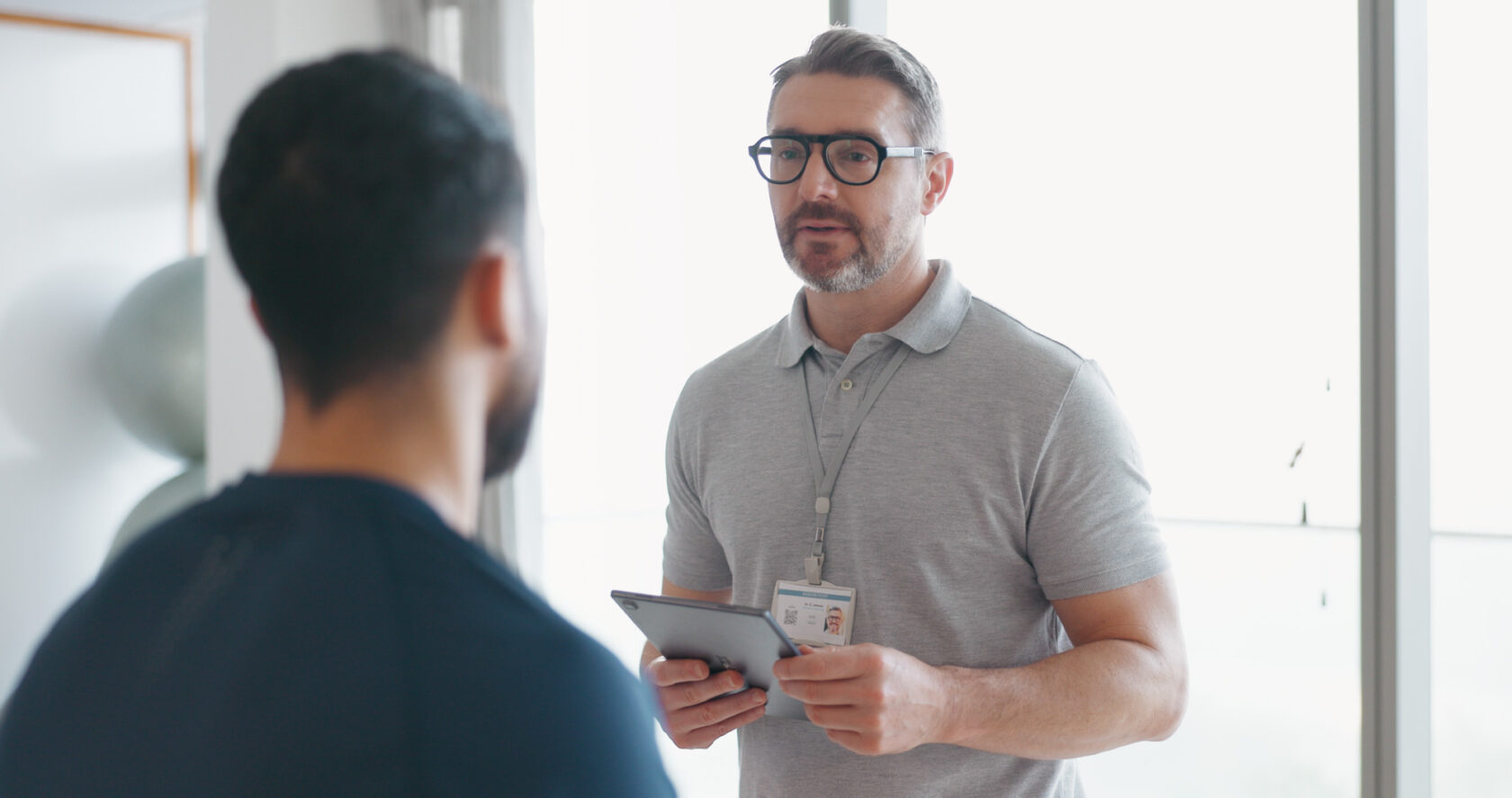 A man in a gray polo shirt with an ID badge holds a tablet and talks to another man facing away from the camera.