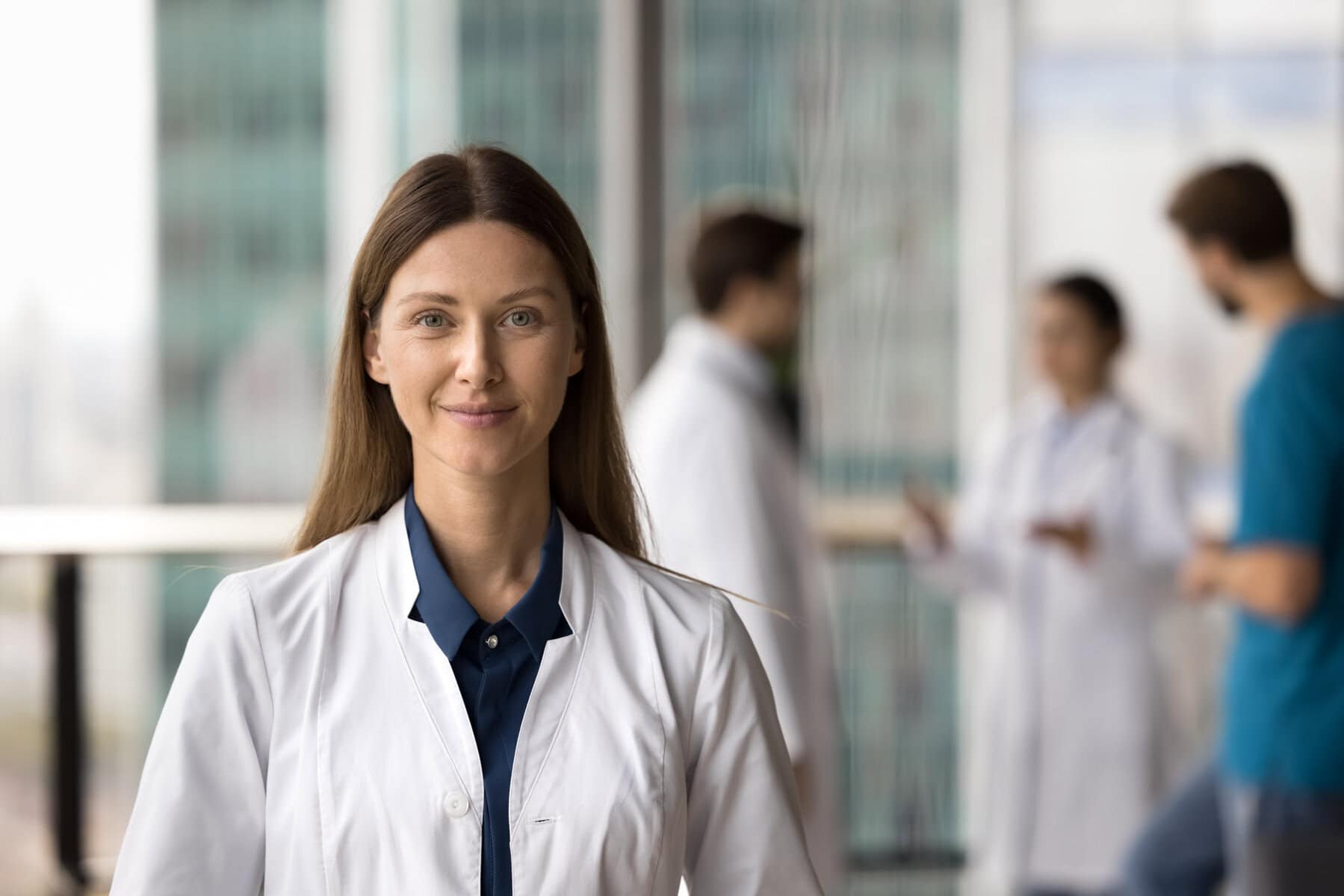A woman in a white lab coat stands smiling in the foreground, while three people in similar attire converse in the blurred background of a modern, light-filled building.
