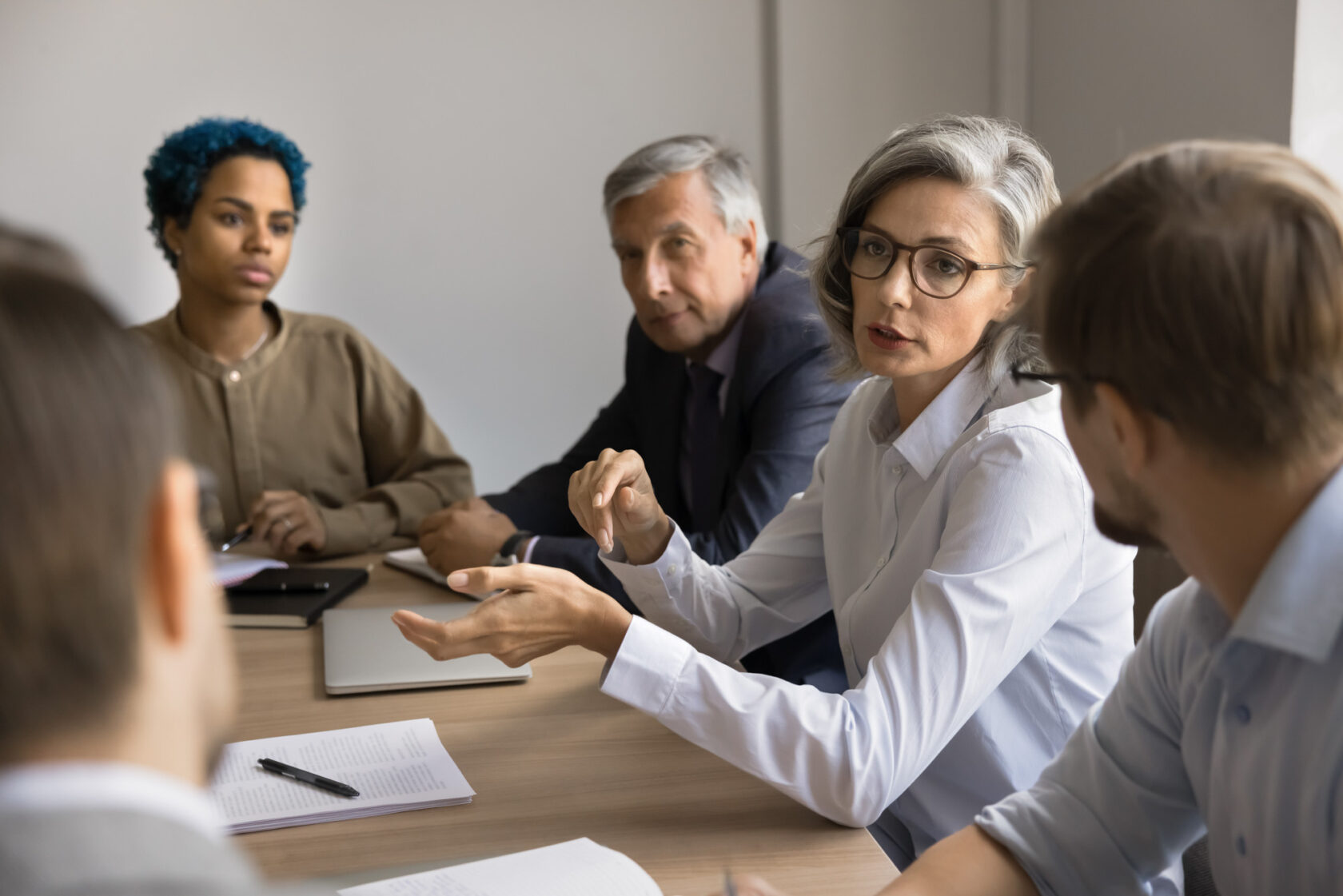 A group of people in a meeting. A woman with gray hair and glasses speaks, while others listen. Notebooks and pens are on the table.