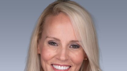 A woman with long blonde hair smiles at the camera against a gradient gray background.