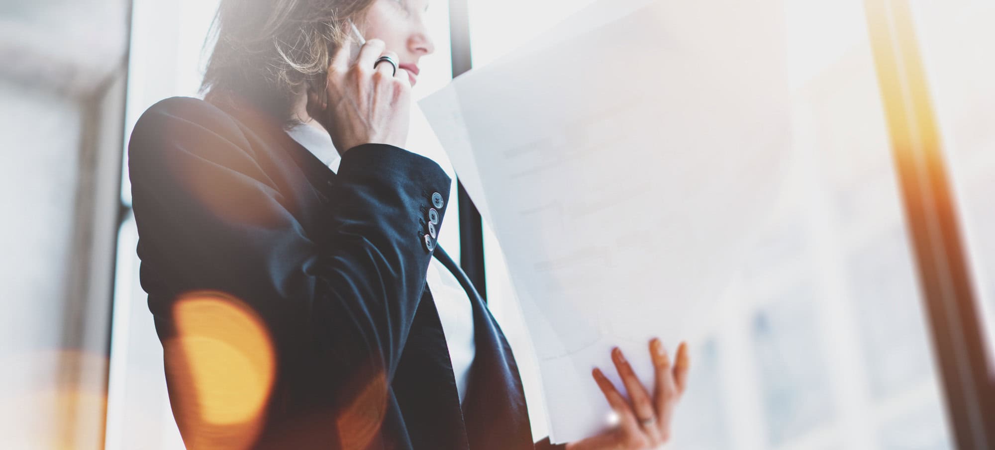 A person in a suit, embodying professionalism, uses a phone while holding papers, standing near a window with bright light. Their poised demeanor suggests they're coordinating appointments for a mental health clinic.