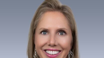 A woman with blonde hair smiles, wearing a black top and silver earrings, photographed against a gradient grey background.