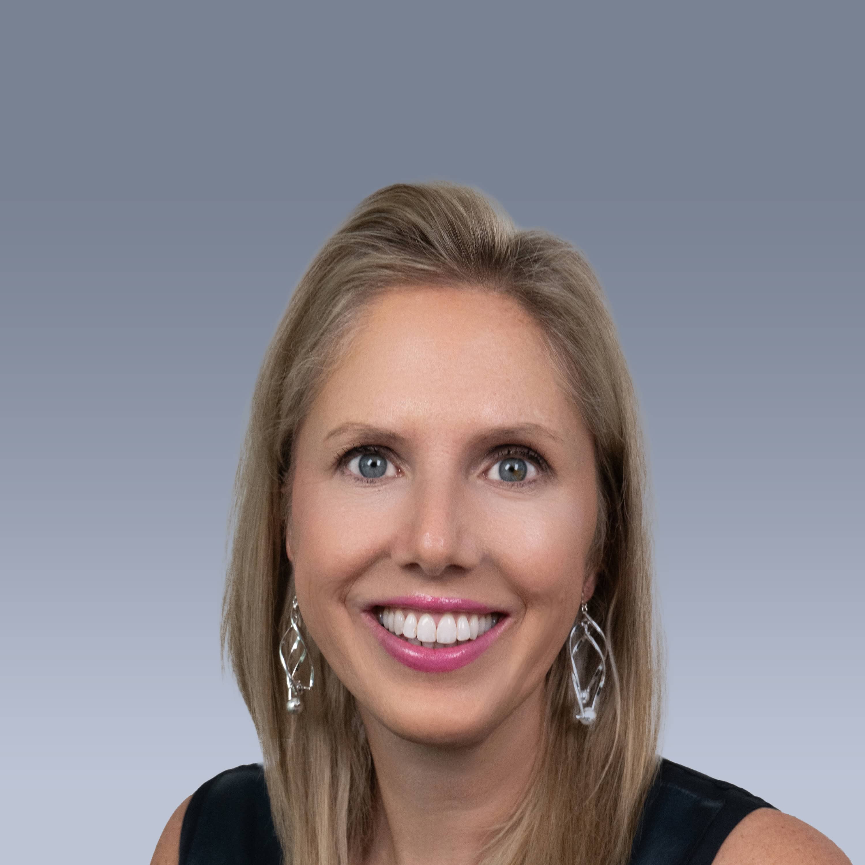 A woman with blonde hair smiles, wearing a black top and silver earrings, photographed against a gradient grey background.