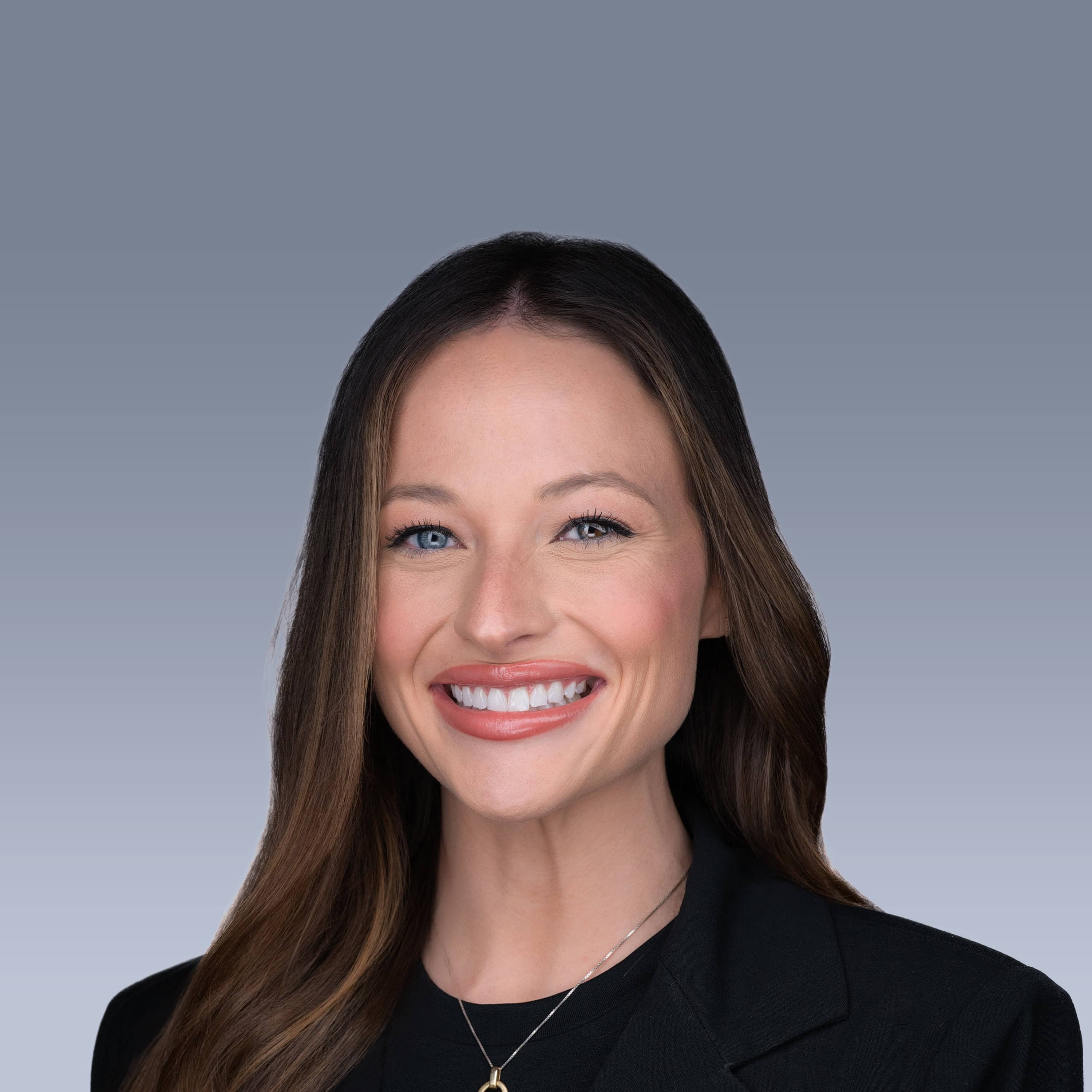 A person with long, dark hair in a formal black outfit smiles in front of a plain grey background.