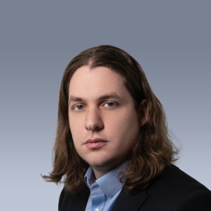 A person with long brown hair, wearing a light blue shirt and dark blazer, is looking towards the camera with a neutral expression against a gray background.