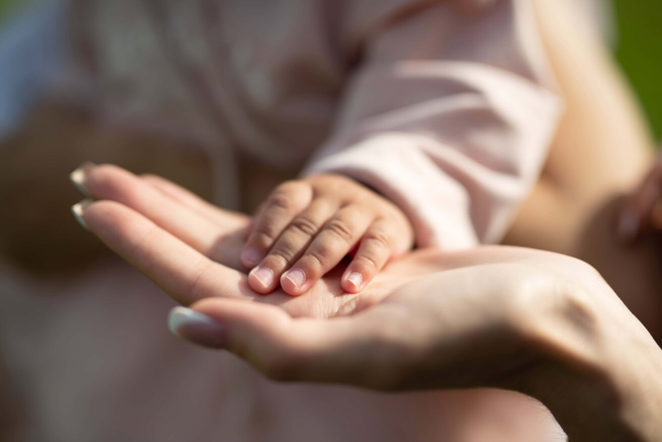 A close-up of an adult's hand holding a baby's hand. Both hands are shown palms up, with the baby's hand resting gently on the adult's palm.