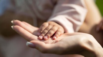 A close-up of an adult's hand holding a baby's hand. Both hands are shown palms up, with the baby's hand resting gently on the adult's palm.