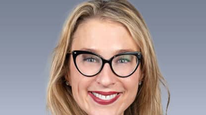A person with long blonde hair, wearing glasses and a black outfit, smiles in front of a gray gradient background.