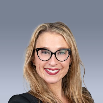 A person with long blonde hair, wearing glasses and a black outfit, smiles in front of a gray gradient background.