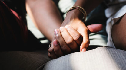 Two people holding hands while seated, with sunlight partially illuminating their hands. They wear casual clothing and one wears a bracelet.