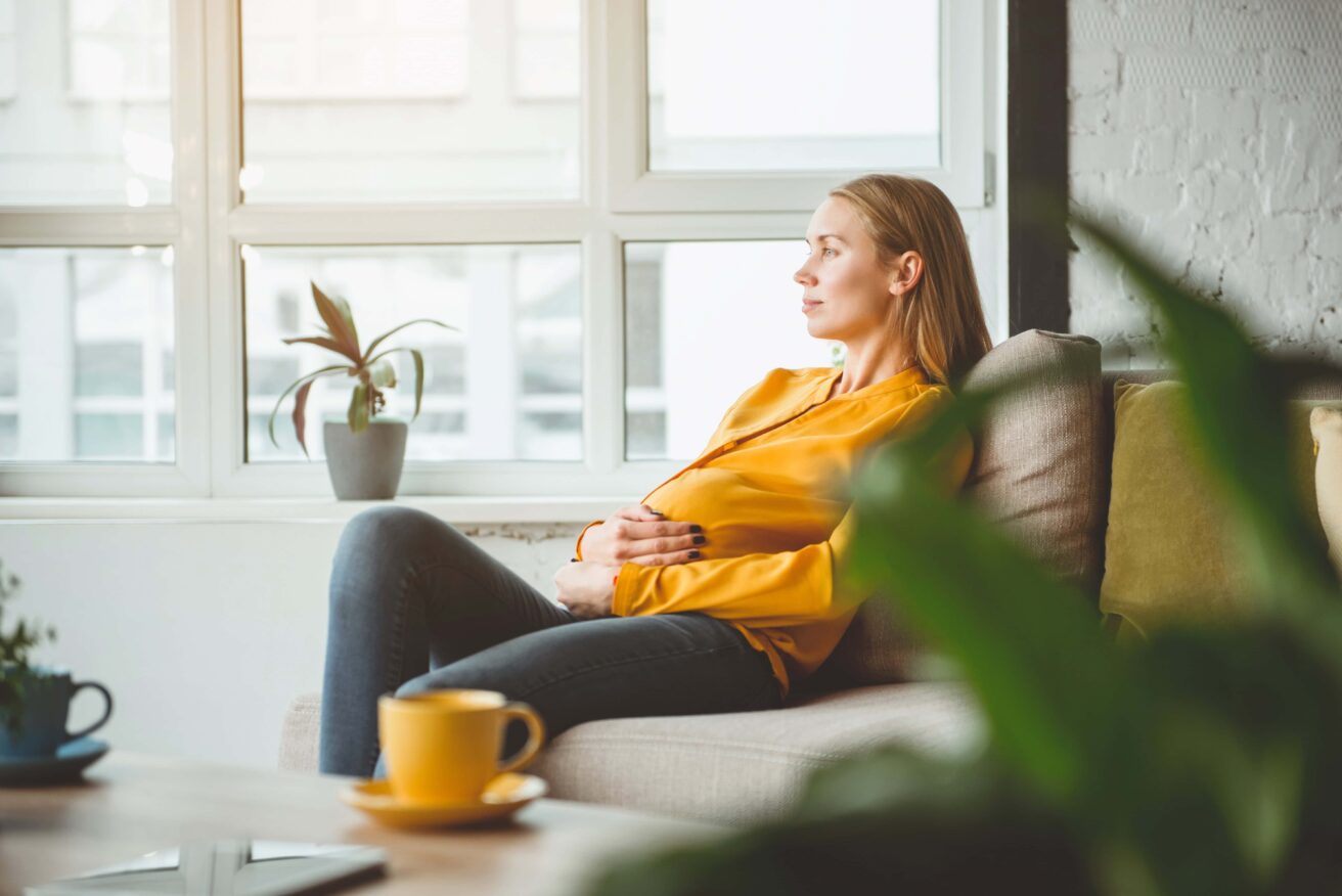 Woman in a yellow sweater sitting on a couch, looking out a window. A small plant is on the windowsill, and a yellow mug is on the table in the foreground.