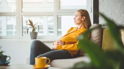 Woman in a yellow sweater sitting on a couch, looking out a window. A small plant is on the windowsill, and a yellow mug is on the table in the foreground.