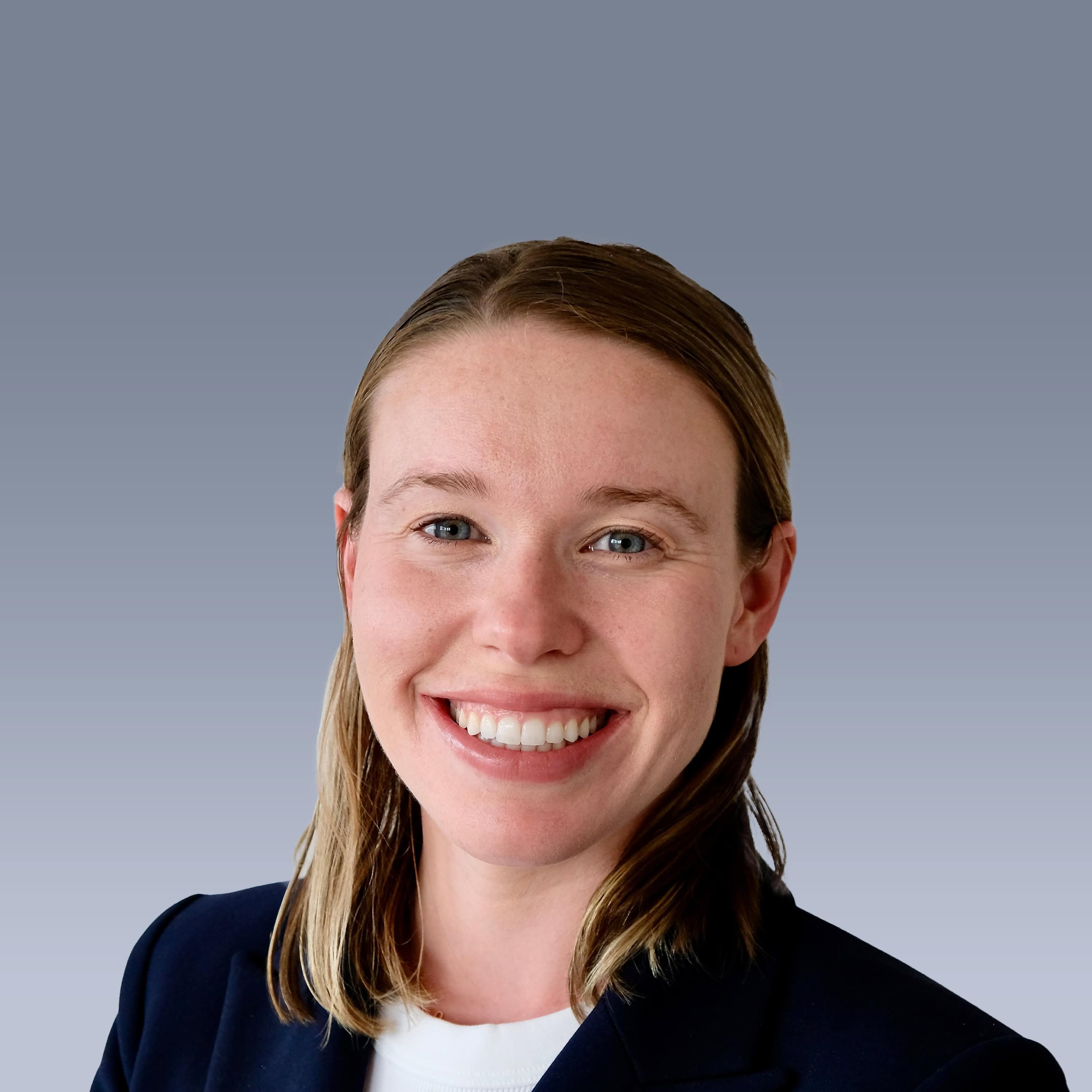 Person smiling in front of a gray gradient background, wearing a dark blazer and white shirt.