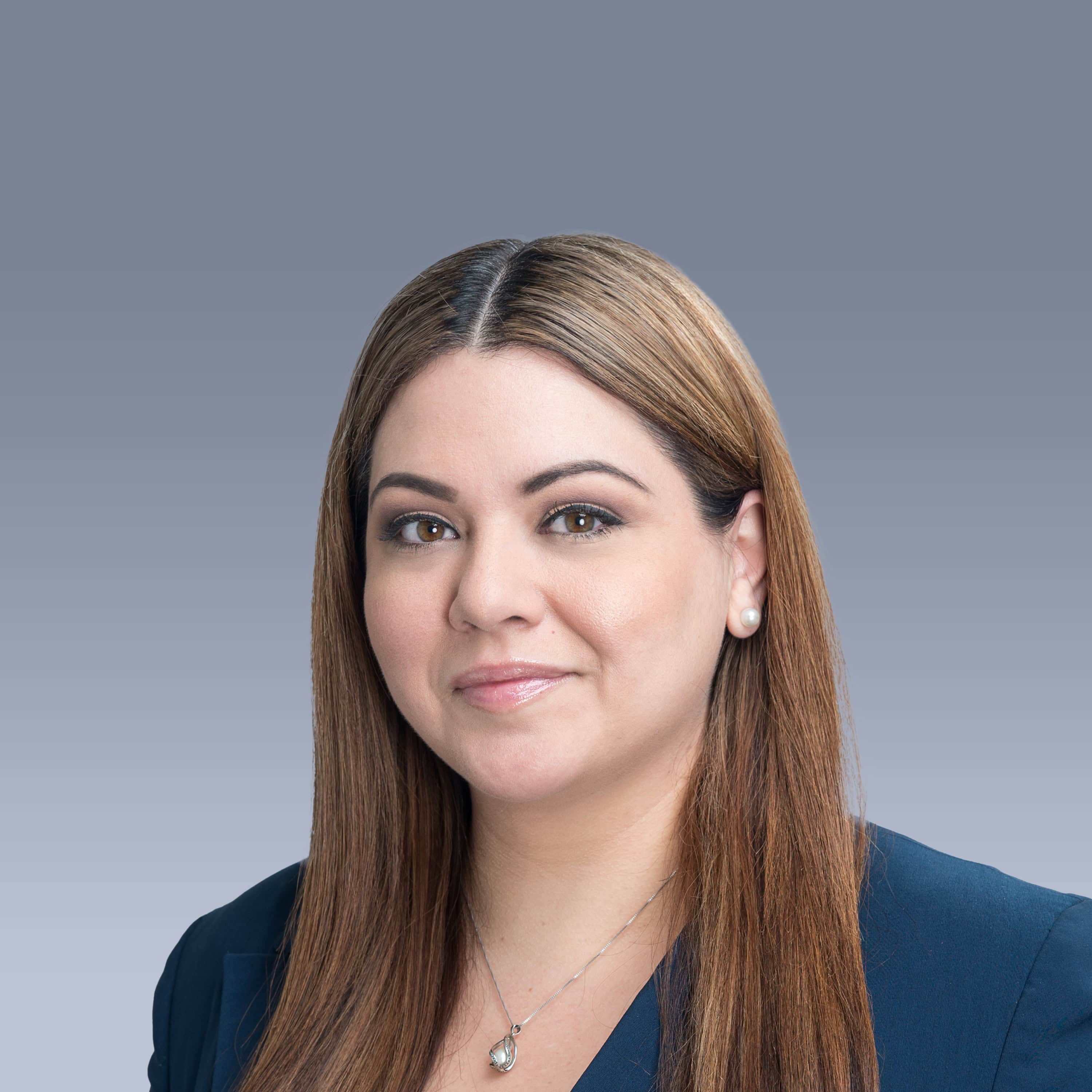 Portrait of a person with long brown hair wearing a navy blazer, a light necklace, and pearl earrings, against a gray background.