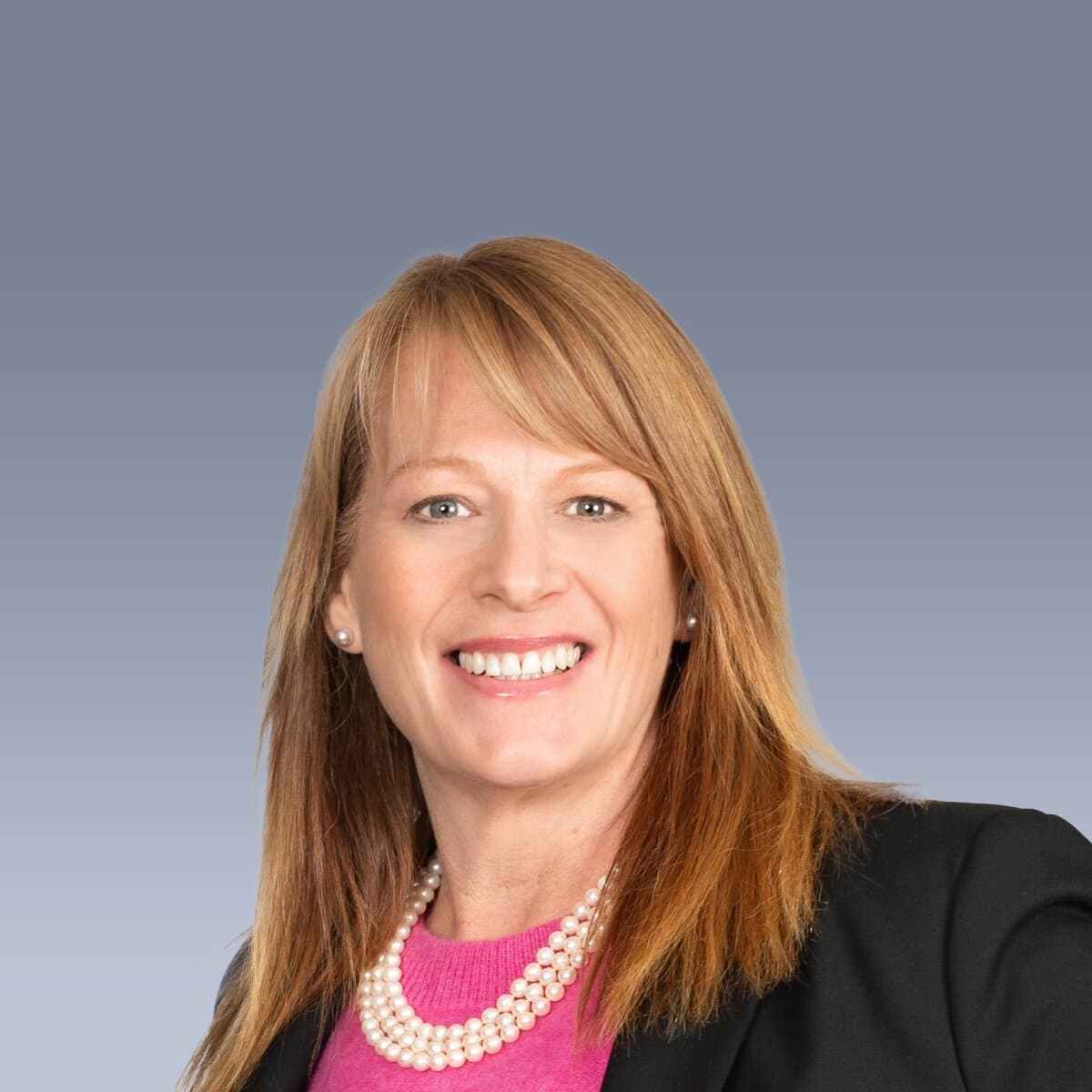 A woman with straight, shoulder-length hair smiles. She is wearing a black blazer, pink top, and a pearl necklace against a gray background.