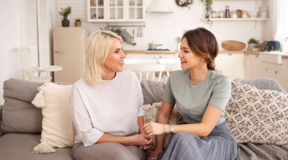 Beautiful happy mature mother and her daughter spending time together after long separation, sitting in cozy living room interior and talking, looking at each other and smiling